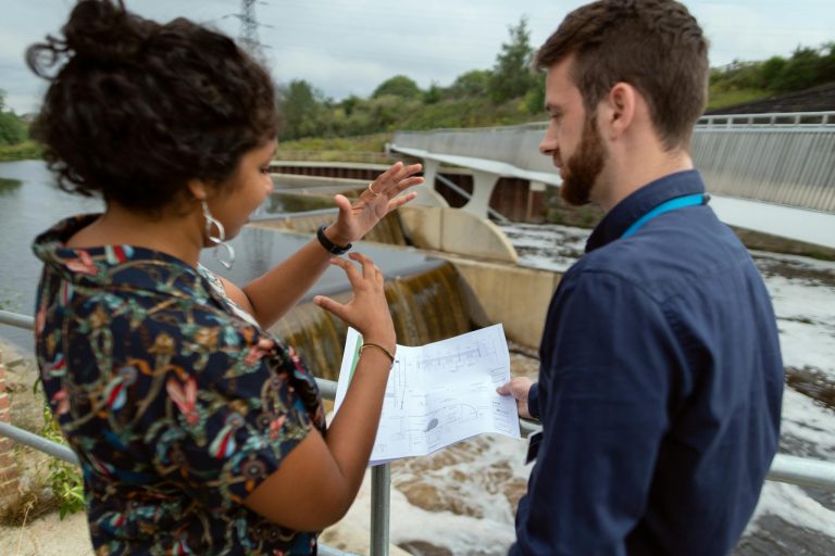 Two engineers discussing plans by a water project site. Collaborative teamwork on hydropower.